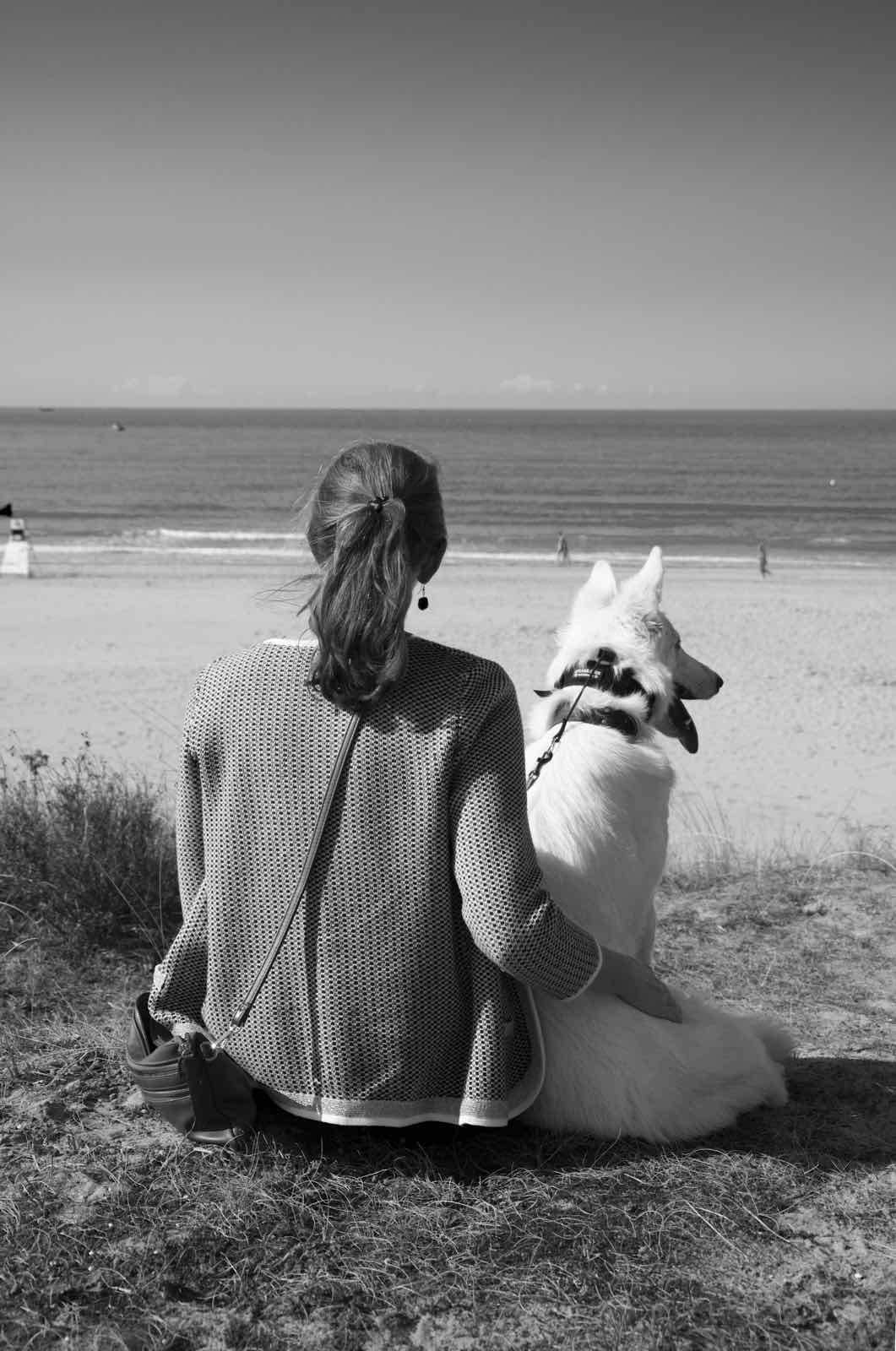 Annelies zit op het strand naast haar witte hond en kijkt naar zee